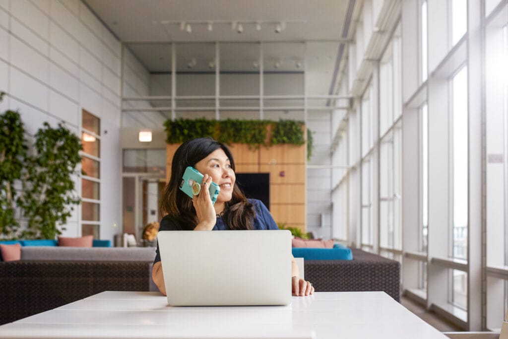 A woman sits at her laptop while on the phone, smiling out the window