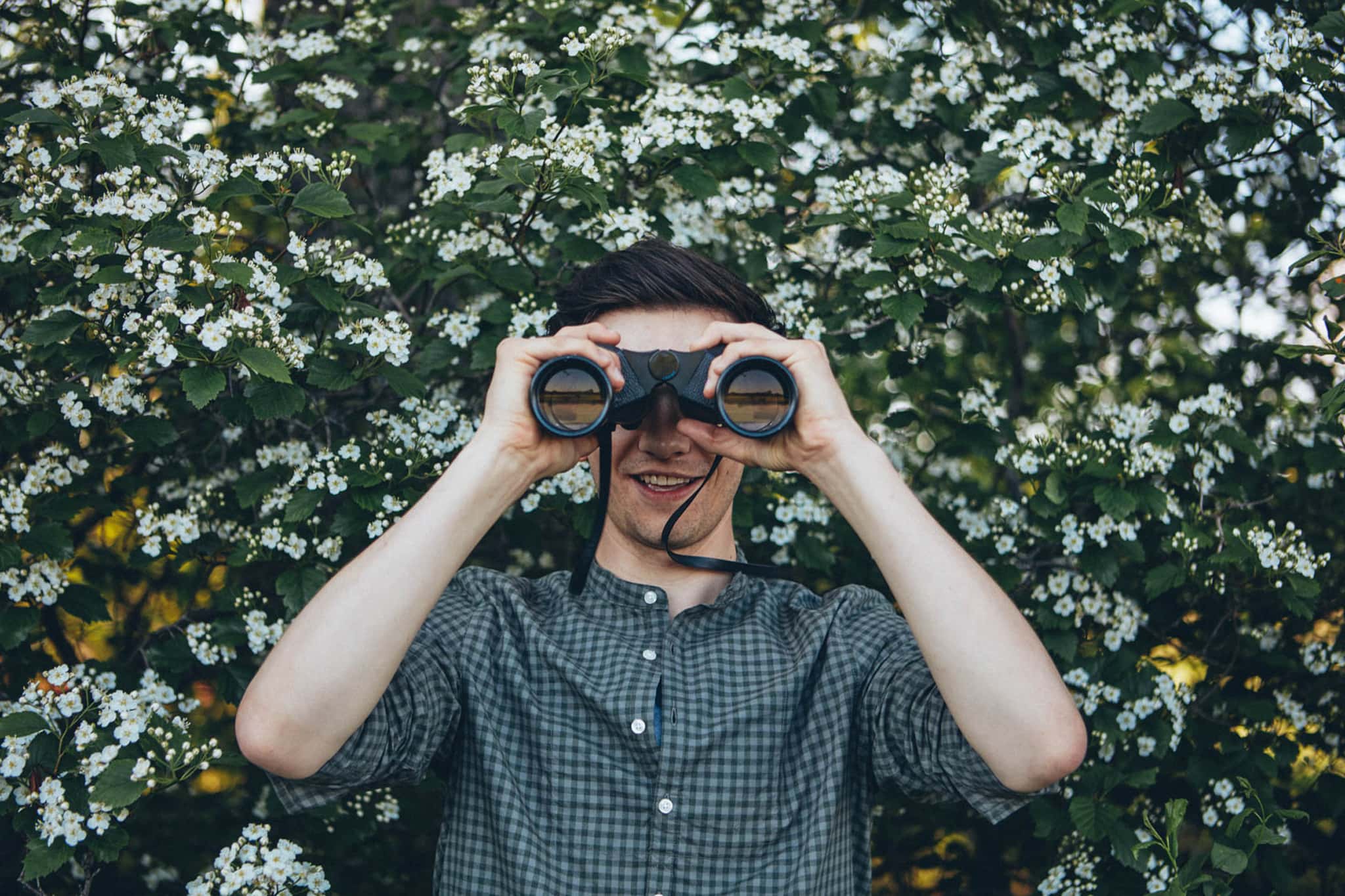 man looking into binoculars in front of flowering shrub